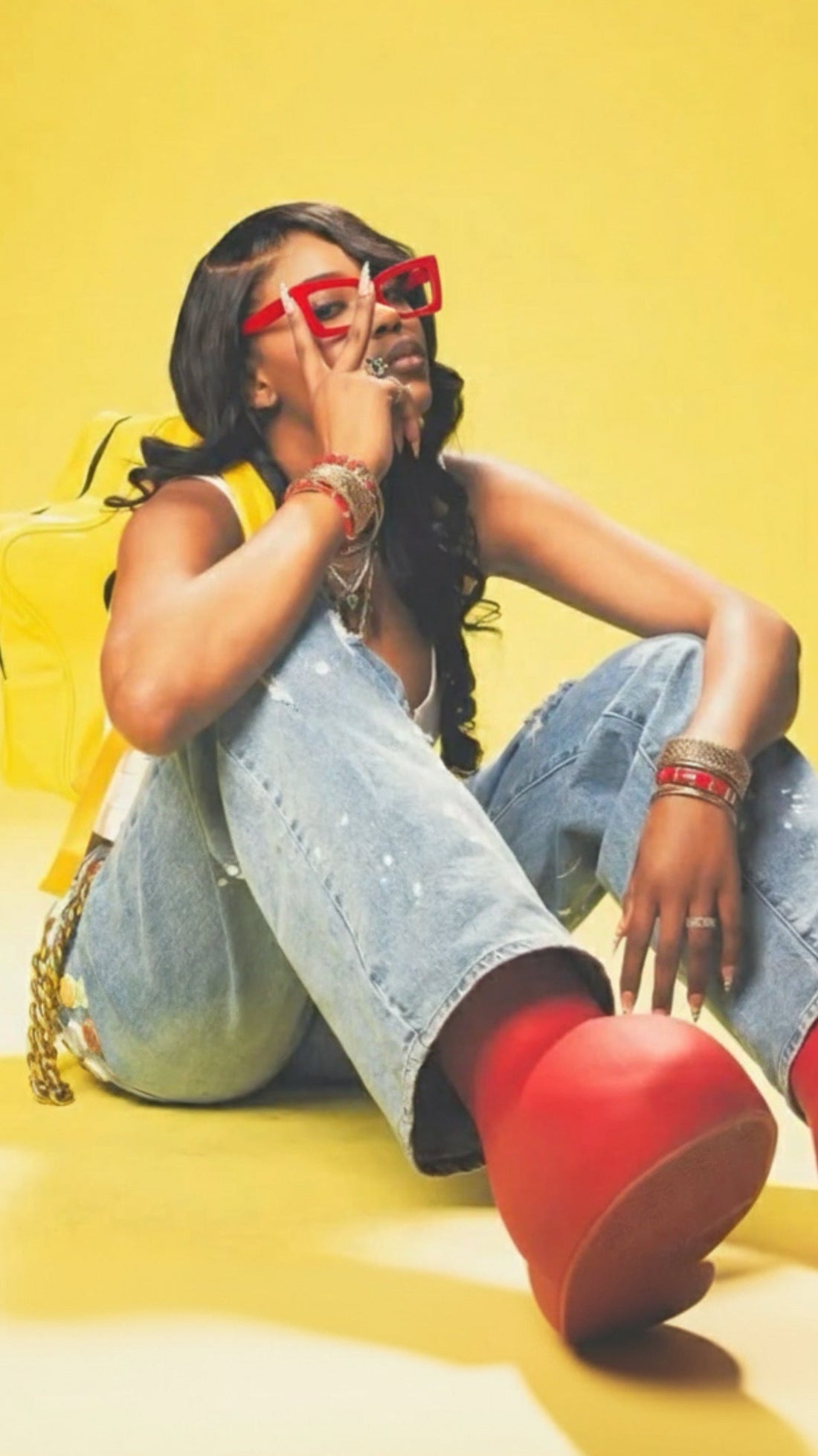 Woman sitting on a yellow floor wearing red shoes, denim jeans, and red sunglasses with a yellow background.