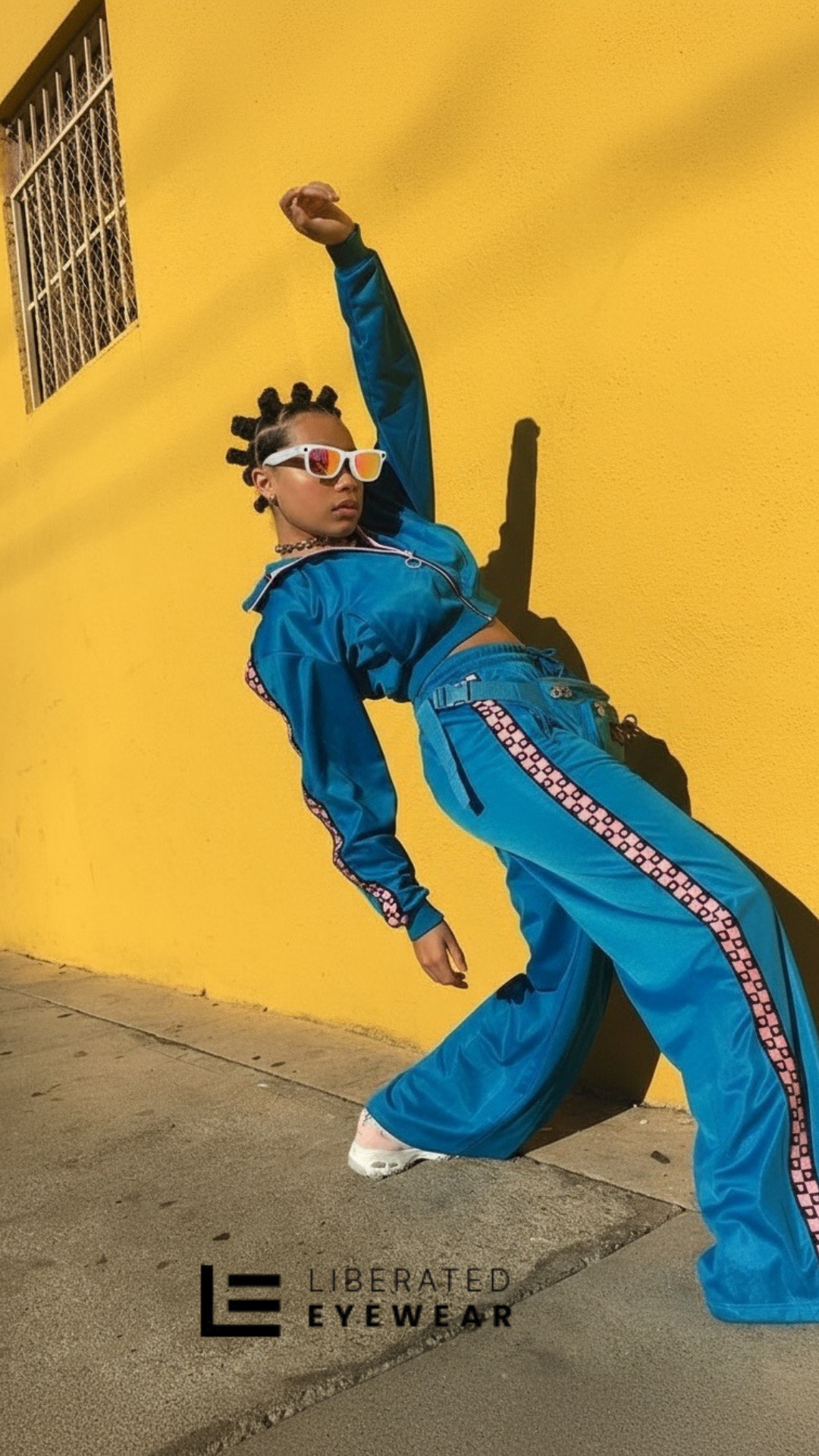 Person in a blue tracksuit with sunglasses against a yellow wall, Liberated Eyewear logo visible.