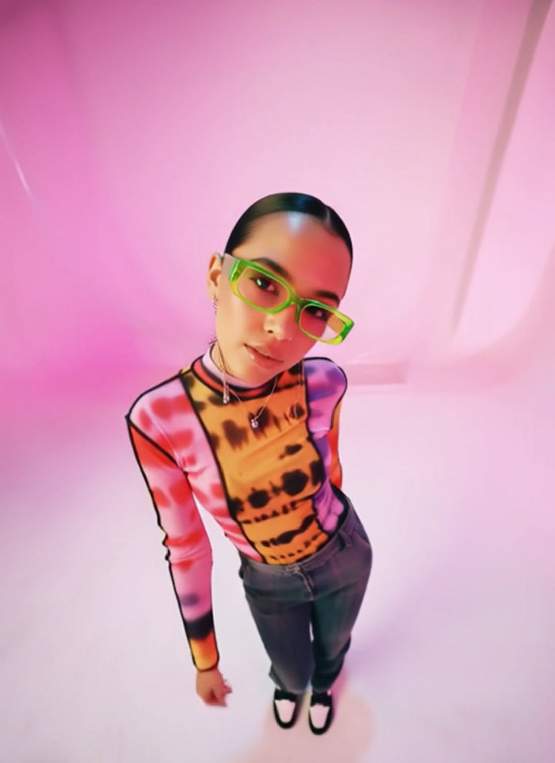 Person wearing a colorful sweater and green glasses against a pink background
