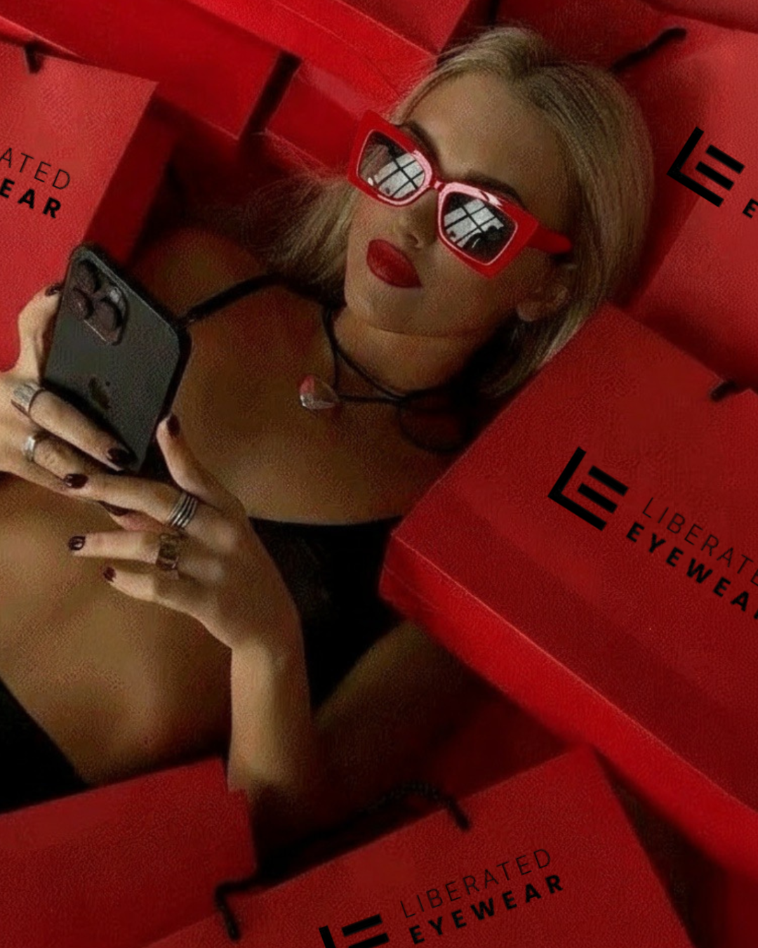 Person wearing red sunglasses and a black bikini lying on red boxes with 'Liberated Eyewear' branding.