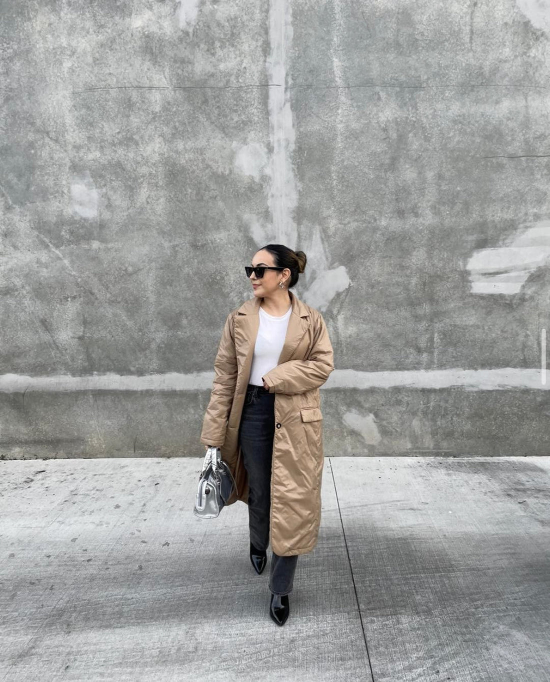 Woman in a beige trench coat walking on a city street with a concrete wall in the background