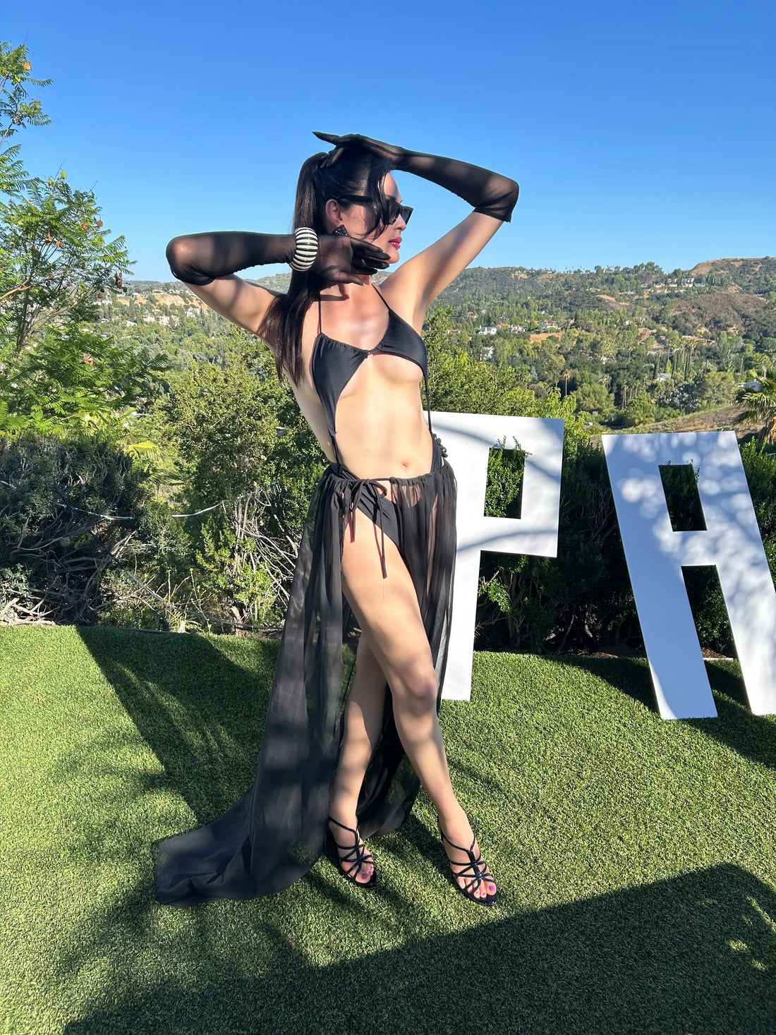 Person in a black outfit standing on grass with trees and a clear blue sky in the background
