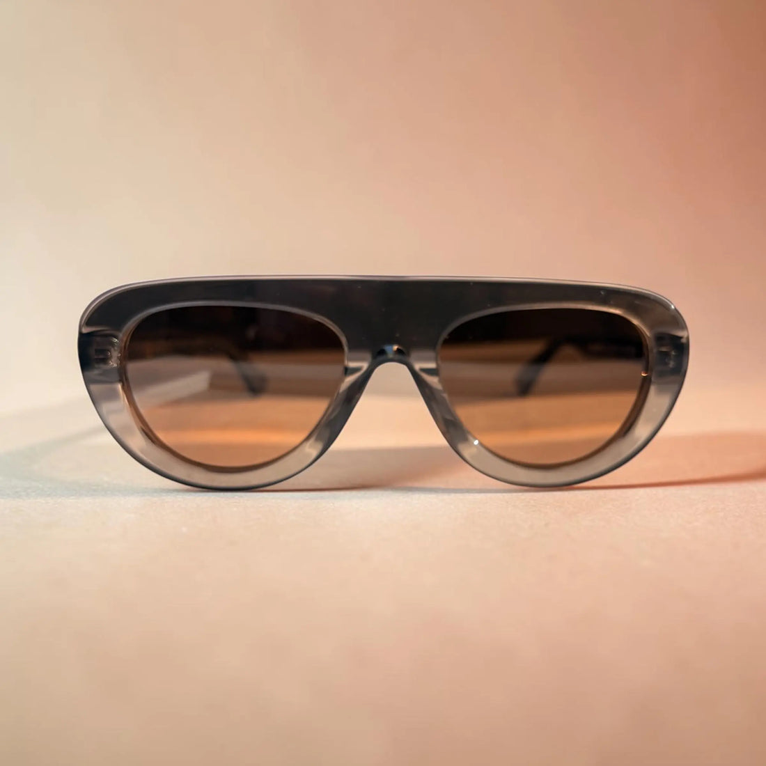A pair of oversized shield-style sunglasses with a translucent dark gray frame and warm brown tinted lenses, photographed against a soft peach-beige background.