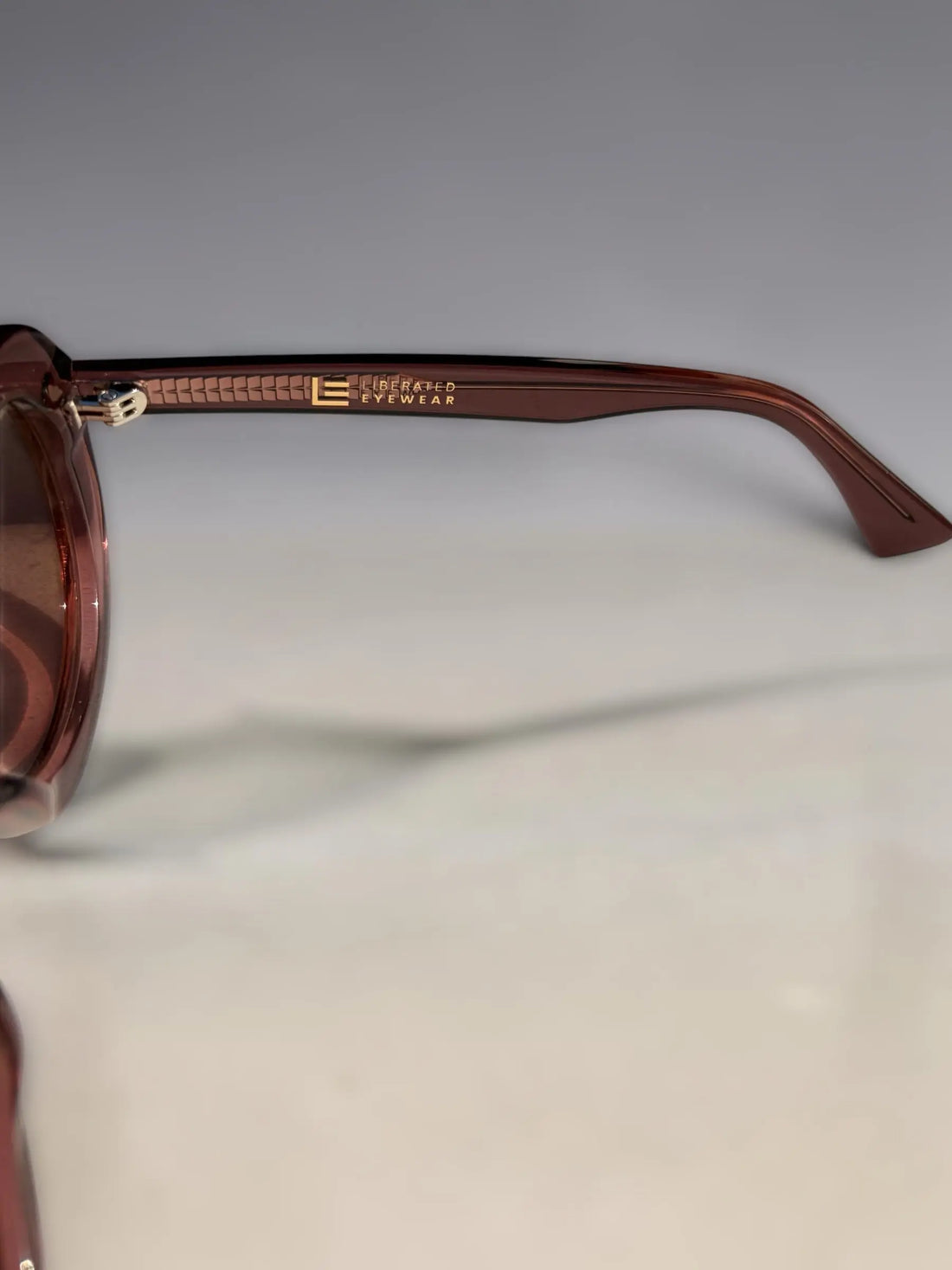 Close-up of a pair of sunglasses with translucent dark pink frames and gold-tone branding on the temple arm, set against a neutral gray background.