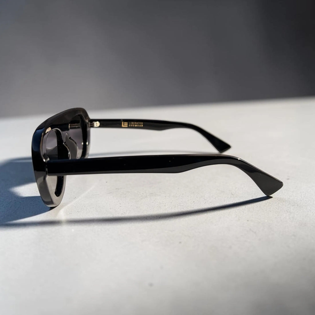 Black acetate sunglasses with a square frame and dark lenses, resting on a light gray surface with a soft shadow. The brand name 'L'OR' is printed in gold on the inner temple arm.