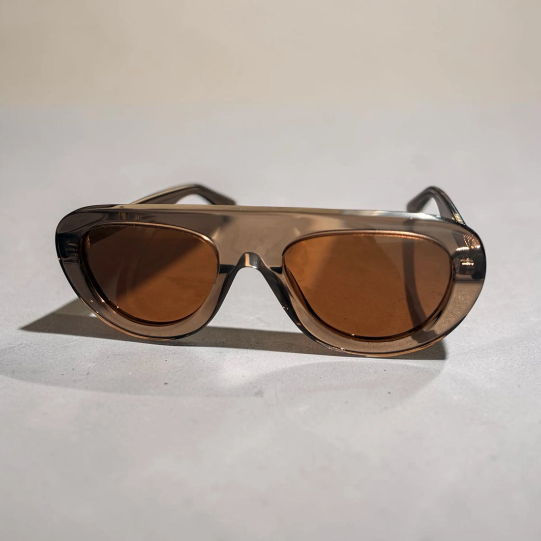 Oversized sunglasses with a translucent smoky brown frame and warm amber-brown lenses, photographed on a light neutral surface.