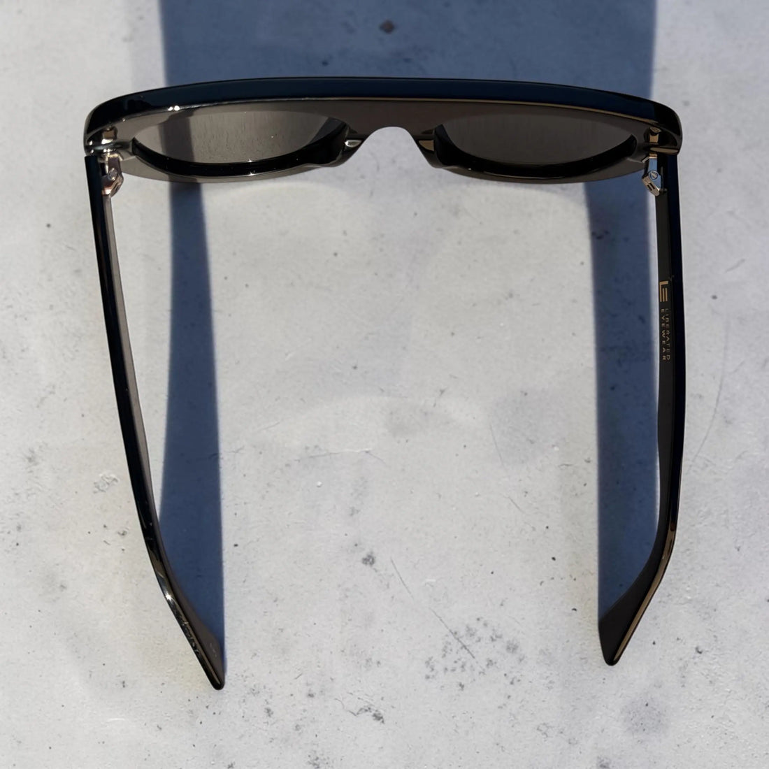 A pair of dark-framed sunglasses with dark lenses, photographed from above on a light gray textured surface with a strong shadow cast to the left.