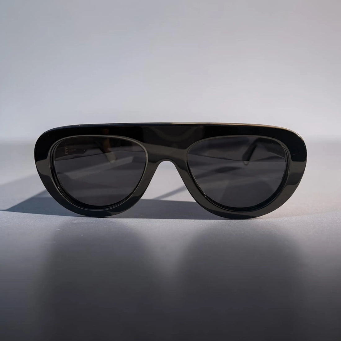 A pair of oversized shield-style sunglasses with a glossy black acetate frame and dark tinted lenses, photographed on a neutral light gray background.